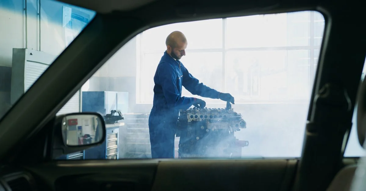 Mécanicien en salopette bleue travaillant sur un moteur dans un garage avec de la vapeur visible autour.