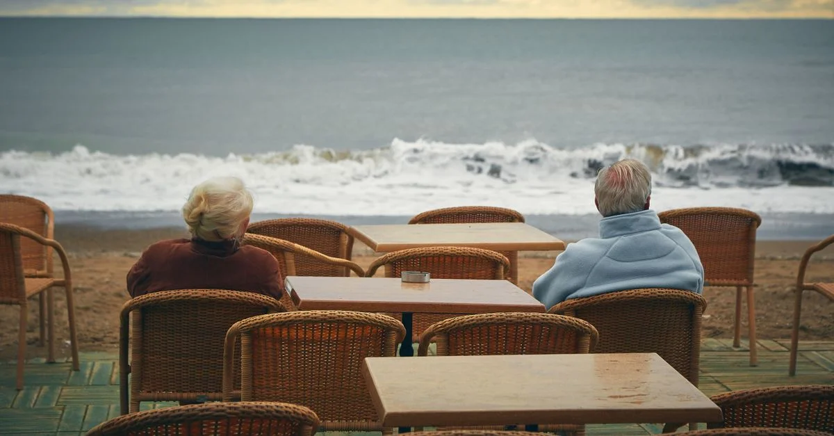 Couple âgé assis dans un café en bord de mer, admirant la vue sur l'océan par une journée nuageuse.