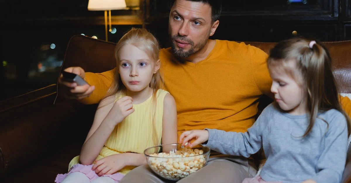 Un père et ses deux filles profitant d'une soirée cinéma avec du popcorn sur le canapé.